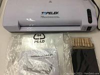 Top view of white Topelek thermal laminator with laminating pouches and wooden clothespins in front