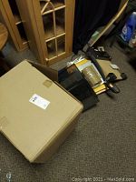 Box with office and shipping supplies including tape, paper cutter, folders, and scale on floor.