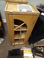 Front view of one light colored wooden cabinet with glass door featuring decorative cross and arc wooden grid pattern. Cabinet has small wooden knob and some surface scuffs.