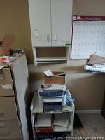 Front view of a wall-mounted double door cabinet above a small white stand holding a printer and office supplies, showing wear and usage marks.