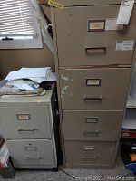 First photo showing both metal file cabinets side by side. The larger cabinet has peeling paint and some dents on the front drawer panels. The smaller cabinet shows scratches and stains on the drawers.