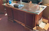 Front view of large wood executive desk with a glass top showing nicks and scratches. Items on desk include a calculator and water bottle.