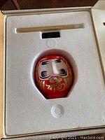Top-down photo of red Daruma doll in foam packaging inside box with wooden paintbrush.