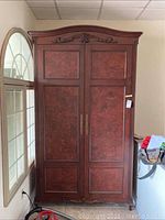Front view of large burled walnut armoire against wall next to arched window, showing double doors with burled wood panels, brass handles, and carved top detail.