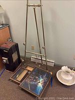 Full view of Stanrite metal easel standing next to several framed vintage wall art prints on the floor and a white decorative ceramic piece.