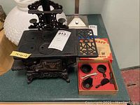 Front and side view of Crescent miniature cast iron stove showing embossed brand and ornamental details, with set of mini cast iron pots and pans in box nearby.