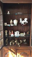 Full cabinet display of a variety of figurines, vases, carousel horses, and small decorative items arranged on glass shelves.