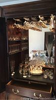 Full view of large glassware lot including glasses hanging overhead, shelves filled with various sizes of etched glasses, tumblers, and glassware on a dark wood cabinet surface with drawers below.