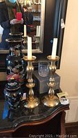 Photo showing two tall brass candle holders, Asian style black vase and ginger jar, and one Mikasa crystal tumbler on a reflective dark wood surface.