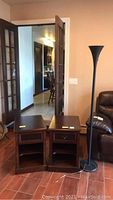 Two wooden side tables and black metal floor lamp placed on floor near a door and brown chair.
