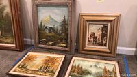 Four framed paintings on floor leaning against wall, one a cabin and mountain scene by Louis M. Cevetello, others include street scene, birch forest, and barn images