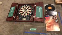 Full view of wooden dartboard cabinet with doors open, electronic scoreboard below, a set of darts, and multiple visible LP records stacked on the floor.