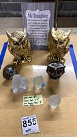 Lot arranged showing two large gold-tone hollow owl figurines front and center with glass owls and small dark owl figurine in front, wooden plaque in background