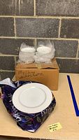 Multiple white Crown Victoria Lovelace china pieces, including plates and smaller plates or saucers wrapped in bubble wrap on a table with handwritten crate label