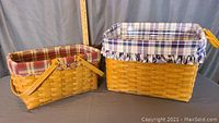 View of two woven baskets with plaid fabric liners and handles, highlighting the medium market basket with red plaid liner and the larger basket with blue/white plaid liner and leather handles.