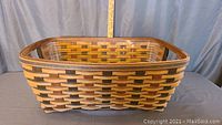 Front view of the small woven wooden laundry basket showing the basket's rectangular shape, woven pattern in light and dark wood strips, and the plastic protector inside the basket.