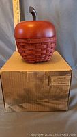 Photo of the Longaberger Red Apple Basket set showing the woven red basket with wooden lid placed on top of the original box with a measuring ruler showing approximate dimensions.
