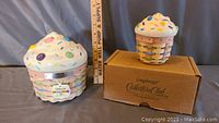 Two pastel-colored woven cupcake baskets with ceramic sprinkle-decorated lids; size difference visible with ruler; smaller cupcake in original box