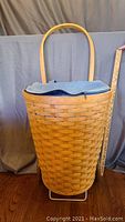 Front view of tall woven Longaberger laundry basket with denim liner and wooden handle. Shows basket's height and width around 24" high and 13" wide.