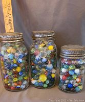 Three clear antique glass jars filled with colorful vintage marbles, two with Kilner metal lids, one with Foresters Glass lid, ruler for scale