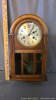 Full view of antique wooden wall clock with rounded top and glass front panel showing pendulum inside. Clock measures roughly 18 by 10 by 5 inches.