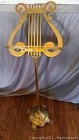 Photo of full antique brass music stand showing lyre-shaped holder and ornate base on wooden floor with grey backdrop.