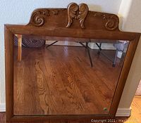 Full view of rectangular oak framed mirror standing on floor against wall. Decorative carved detail visible on top center of frame. Mirror reflects hardwood floor and parts of furniture.