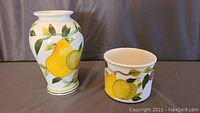 Photo showing the Summer Fruit vase and crock side by side on a grey surface with a dark background.