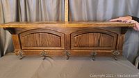 Front view of antique solid wood wall shelf with five ornate metal hat hooks attached along the bottom; shelf has arched panel front.