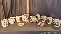 Overview of 8 mugs, 2 small plates, and creamer with saucer in Pomona pattern with fruit decoration on white porcelain.