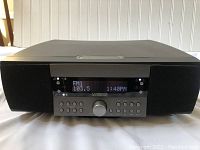 Front view of Cambridge Soundworks Model 735 radio unit showing digital display indicating FM1 103.5 and time 1:40 PM, central volume knob, and buttons for presets, alarm, and tuning.