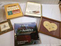 Photo showing three hardcover books and two framed handmade artworks on a white surface.