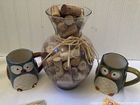Large clear glass vase filled with wine corks tied with twine bow, two owl mugs on each side, and memo pads below