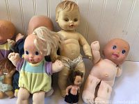 Photo shows seven vintage baby and toddler dolls arranged in a cluster, sitting and leaning against a white panel background. Dolls vary in size, hair style, and clothing state.