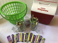 Red and white Igloo Little Playmate cooler, green plastic laundry basket, two vintage Coca-Cola glass mugs with bent handles, and many Crystal Light powdered drink packets arranged on white surface.