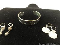Photo of sterling silver cuff bracelet with open back, floral engraving, and two pairs of silver tone earrings, one with textured beaded dangles, the other with flat disk dangles, all on black background.