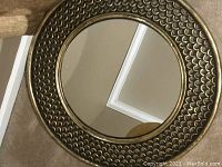 Close up photo of front round mirror showing gold metal tone scalloped textured frame and reflective mirror surface.