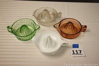 Four vintage glass juicers in green, orange, clear, and milk glass arranged on a white surface showing tops and handles