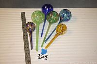 Six blown spatter glass watering orbs shown together on striped fabric background with measuring ruler beside them for scale.