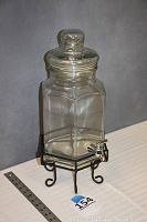 Hexagonal clear glass beverage decanter with clear glass lid and silver-tone spigot sitting on black wrought iron stand with curled feet and hexagonal base holding the decanter. A ruler is placed next to the stand showing approximately 18 inches height.