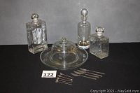 Full set of glass decanters, cheese dome with plate, five appetizer forks, and four appetizer knives arranged on black cloth background.