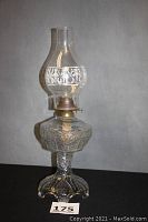 Full view of antique oil lamp standing on a black surface with a grey background. Lamp has a glass base and chimney with decorative elements and a brass collar at the top of the font.
