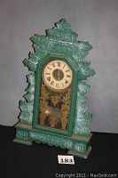 Full front view of the ornate green painted oak mantel clock with carved leaf motifs and gold floral glass design.