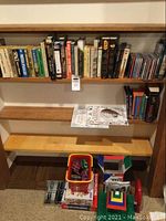 Shelf with various books by different authors and CDs on right side, below shelf holds small bucket with Magformers magnetic toy pieces and other toys
