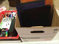 Photo showing black office binders in a cardboard box along with a basket with various markers and a bottle of glue.