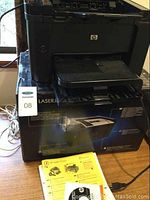 Front angle view of black HP LaserJet Pro P1606dn printer on its box, power cord attached, manual on table.