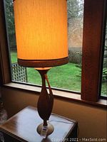 Full view of tall vintage table lamp with lit fabric shade on a wooden table by window.