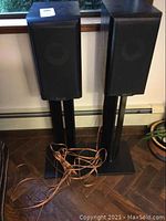 Pair of Mission bookshelf speakers on black metal stands with speaker cables coiled at the base. Speakers are black with removable fabric grilles.