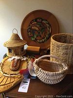 Photo showing multiple items including the wooden plate with Indigenous motif, woven baskets, carved wooden paddle, leather feather decorated item, and small wooden boxes arranged on a surface.