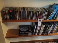 Media collection on two wooden shelves, top shelf with mostly CDs and some DVDs and Blu-Rays, bottom shelf with DVDs and boxed set titled Space Shuttle DVD
